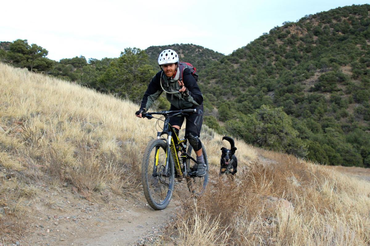 A mountain biker riding on a dirt trail through a grassy area, with a black dog running alongside. The background features rolling hills and trees. The biker is smiling and wearing a helmet and protective gear. North Backbone mountain bike trail.