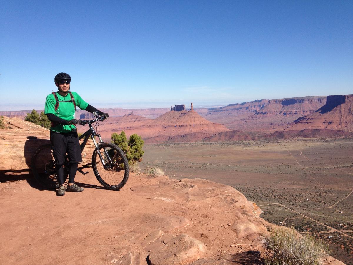 Santa Cruz Tallboy LT: A person wearing a green shirt and cycling gear stands next to a mountain bike on a rocky outcrop, overlooking a vast desert landscape with red rock formations and blue skies in the background.