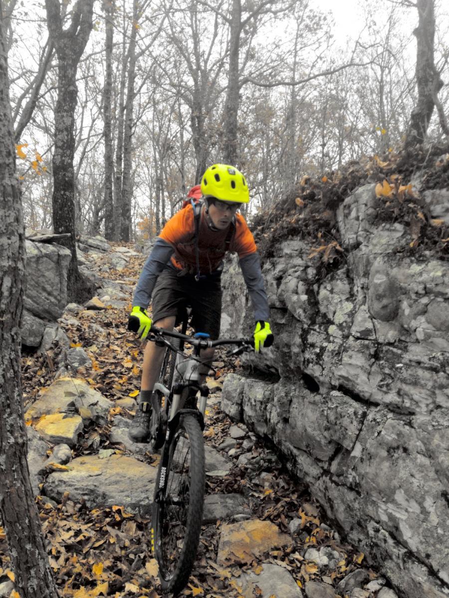 A young person riding a mountain bike on a rocky trail surrounded by trees. They are wearing a bright yellow helmet and neon gloves, with an orange shirt and dark shorts. The trail is covered in fallen leaves and rocks, and the atmosphere appears misty and subdued. Coldwater Mountain mountain bike trail.
