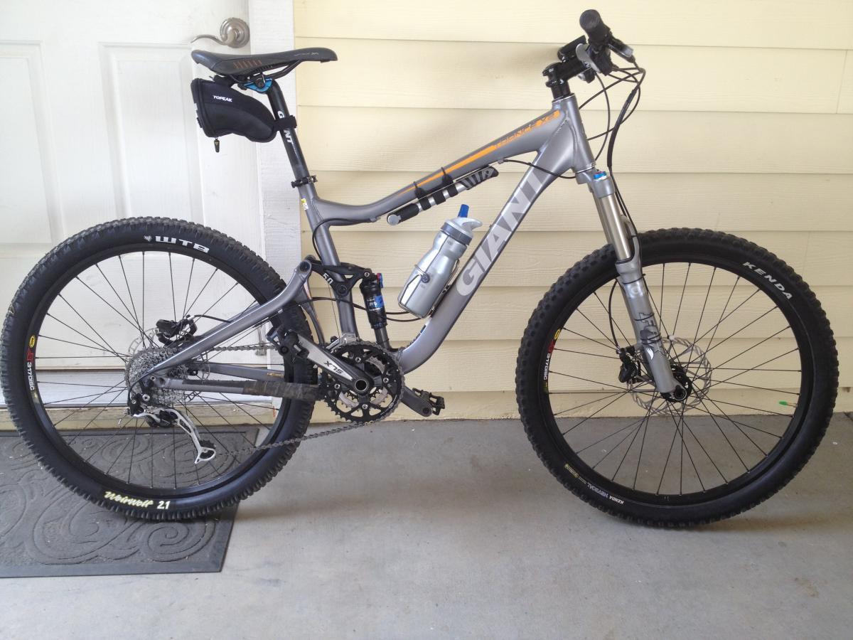 Giant Trance X2: A gray mountain bike with the brand name "Giant" prominently displayed on the frame, positioned next to a white door. The bike features thick tires suitable for rough terrain, a water bottle mounted on the frame, and a small saddle bag attached to the seat. The background includes a textured floor mat and light yellow walls.