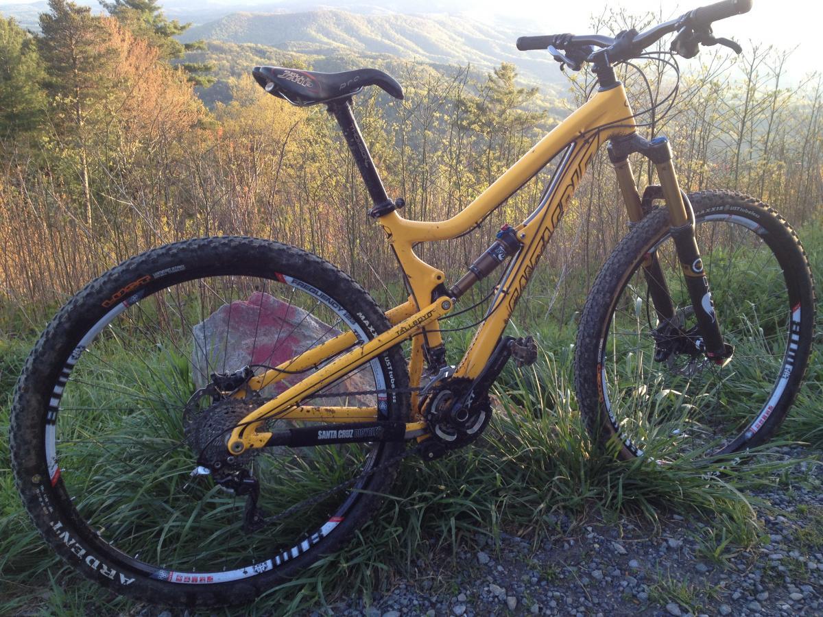 Santa Cruz Tallboy LT: A mountain bike with a vibrant yellow frame is positioned on grass and gravel, with a scenic view of rolling hills and trees in the background, illuminated by soft sunlight. The bike features thick tires designed for off-road terrain and visible components, indicating its suitability for rugged trails.