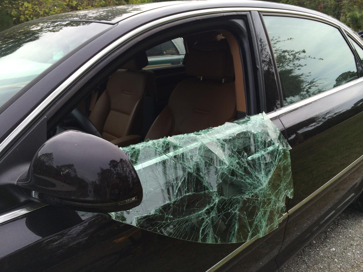 A black car with a shattered driver's side window, covered with jagged pieces of glass. The interior is visible, showcasing beige seats. The car is parked in a natural setting with greenery in the background. Caloosahatchee Regional Park mountain bike trail.