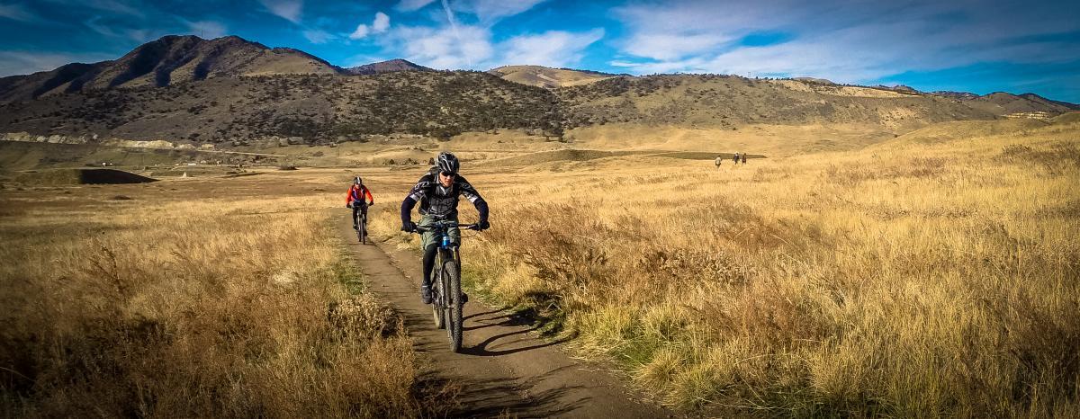 Two mountain bikers riding on a dirt trail through a grassy landscape with rolling hills and mountains in the background under a blue sky with wispy clouds. Green Mountain mountain bike trail.