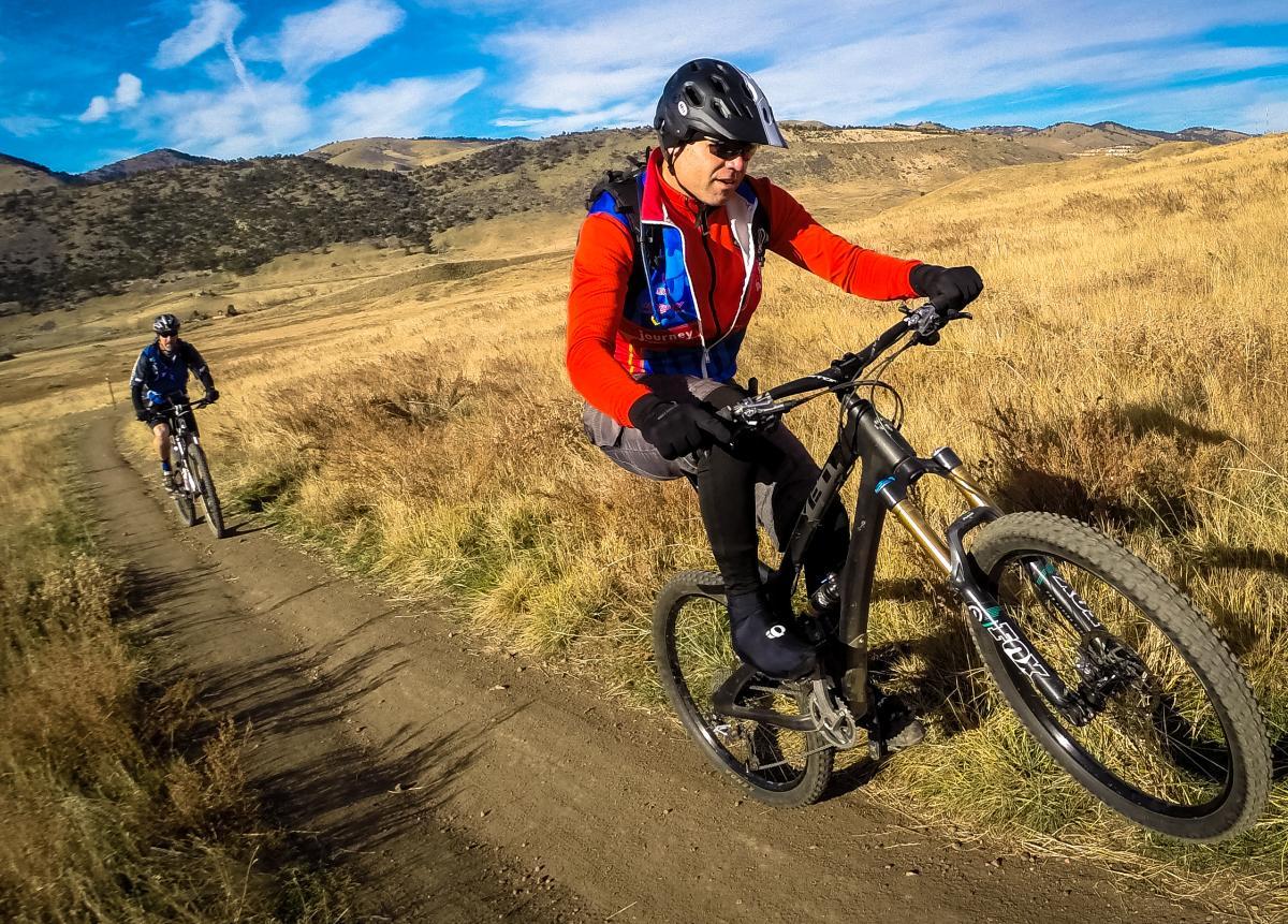 Two mountain bikers riding on a dirt trail through golden grasslands, with hills in the background. One cyclist is in the foreground, wearing a red jacket and a helmet, performing a wheelie, while the other rider follows behind in blue. The scene captures a sunny day with a clear blue sky. Green Mountain mountain bike trail.
