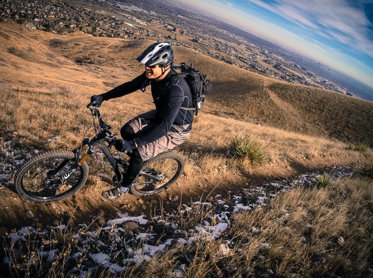 A mountain biker riding on a dirt trail through a dry, grassy hillside with a distant view of a suburban landscape and blue sky. The rider is wearing a helmet and protective gear, navigating the terrain on a black mountain bike. Green Mountain mountain bike trail.