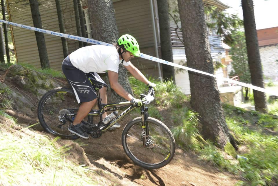 Scott Genius: A person riding a mountain bike down a dirt trail in a forested area. They are wearing a green helmet and a white t-shirt, focused on navigating the terrain. Trees surround the path, and a ribbon marks the course. Sunlight filters through the foliage, highlighting the rider's movement.