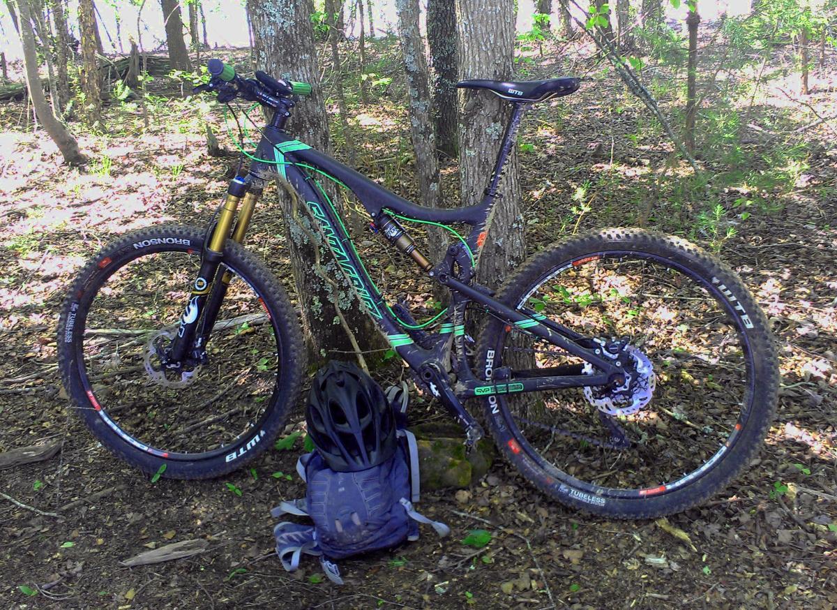 Santa Cruz Blur TRC: A mountain bike with a sleek black and green frame is leaned against a tree in a wooded area. The bike features front and rear suspension, thick tires, and visible components like the handlebars and pedals. Next to the bike, there is a blue backpack and a black helmet on the ground, surrounded by fallen leaves and greenery. Sunlight filters through the trees, creating a natural and serene setting.