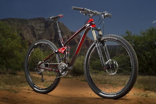 Trek Fuel EX 9.8: Mountain bike standing on a dirt trail surrounded by greenery and rocky terrain, showcasing its sleek design and shiny components. The bike features prominent tires and a vibrant red and black color scheme, with a scenic background of hills and blue sky.