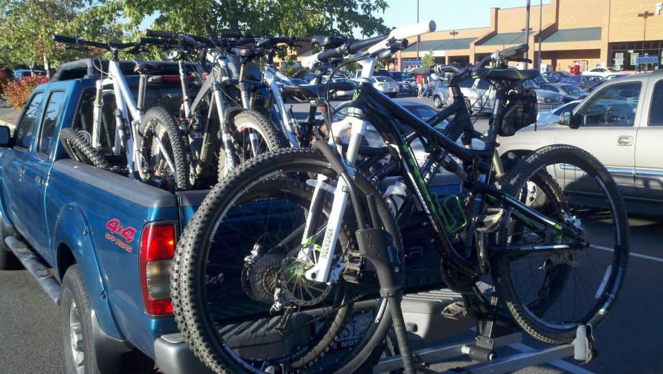 Trek Superfly AL: A blue pickup truck parked in a lot, loaded with several mountain bikes in the truck bed. The bikes are positioned upright, showcasing their tires and frames. In the background, there are other vehicles and a building, suggesting a busy outdoor setting.