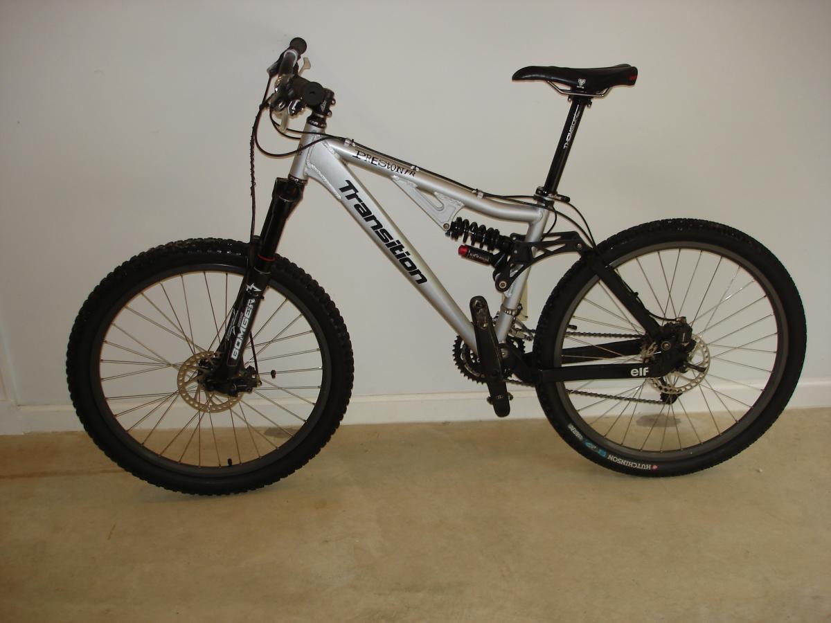 Transition Preston FR: A silver mountain bike with black accents, featuring disc brakes and thick tires, positioned against a plain wall. The bike has a dual suspension design and is designed for off-road riding.