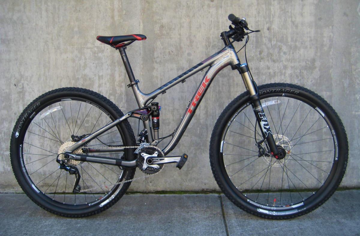 Trek Fuel EX 8: A side view of a silver Trek mountain bike with black tires and red accents, positioned against a concrete wall. The bike features a front suspension fork, disc brakes, and a modern gear system, showcasing its design for off-road cycling.