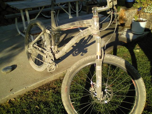 Ellsworth Truth: A dusty mountain bike parked on a concrete surface, with a picnic table and greenery in the background. The bike shows signs of recent use, with dirt and grime covering the frame and wheels.
