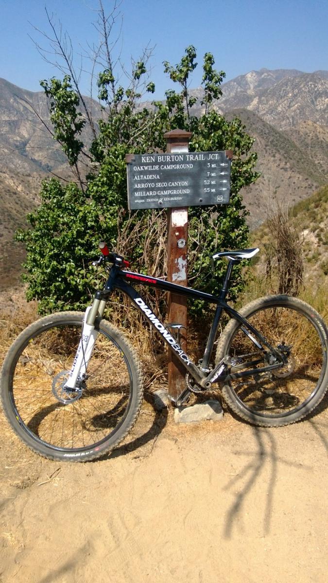 Diamondback Overdrive: A mountain bike leans against a trail sign that indicates the junction for the Ken Burton Trail. The sign lists distances to various locations, including Oakwilde Campground, Altadena, Arroyo Seco Canyon, and Millard Campground. The landscape is dry and mountainous, with sparse vegetation and a clear blue sky in the background.