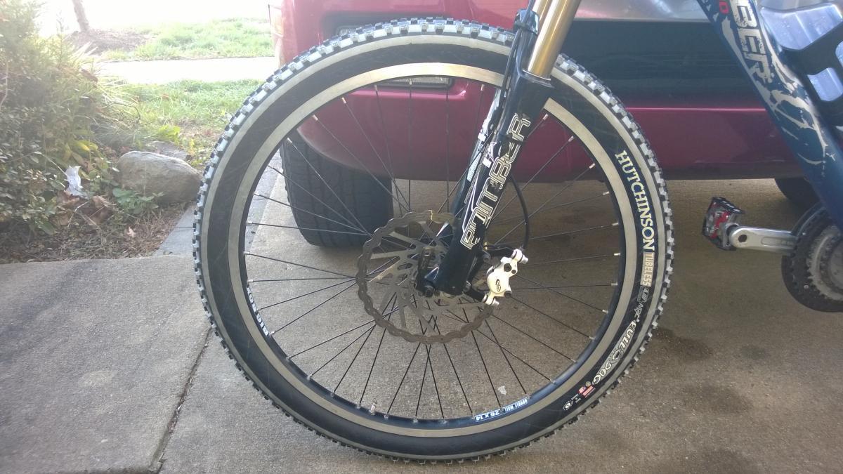 A close-up view of a mountain bike's front wheel, featuring a black tire with a tread pattern suitable for rough terrain. The wheel includes a visible disc brake and is mounted on a fork labeled "PITBER." In the background, a red vehicle is partially visible, along with green grass and a few rocks.