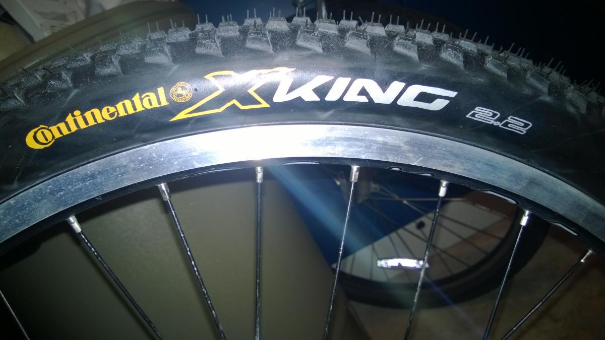 Genesis Sabre V2600: Close-up image of a bicycle tire showcasing the Continental X-King 2.2 model. The tire features a black tread pattern and a silver rim, with prominent yellow and white branding visible.