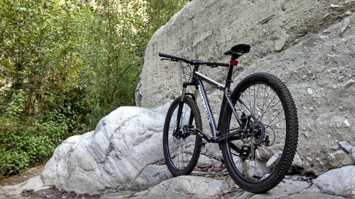 Diamondback Overdrive: A mountain bike resting against a large rock in a forested area, surrounded by greenery and rugged terrain. The bike is black with silver accents and is positioned on a gravel path.