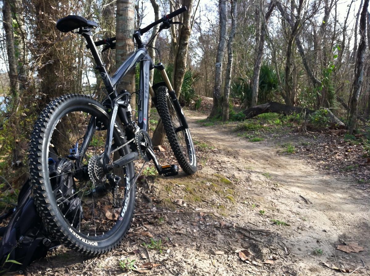 Trek Remedy 7: A mountain bike leaning against a tree on a dirt trail surrounded by trees and underbrush. The path curves ahead, inviting exploration. Leaves are scattered on the ground, indicating a natural outdoor setting.