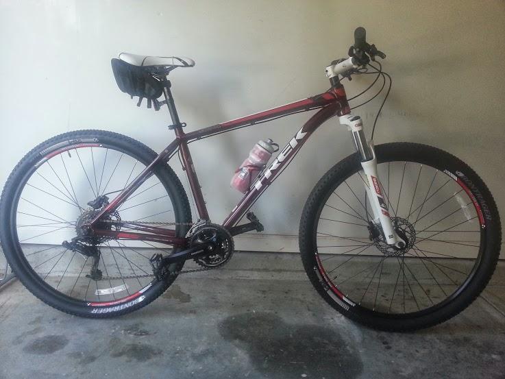 Trek X-Caliber: Mountain bike leaning against a wall in a garage. The bike has a maroon frame with white accents, featuring a suspension fork, wide tires, and a water bottle holder with a bottle. A black saddle bag is attached to the seat. The concrete floor is visible below.
