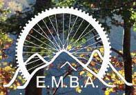 Logo of the E.M.B.A. featuring a stylized bicycle wheel with a mountain silhouette in the center, surrounded by greenery and autumn leaves in the background.