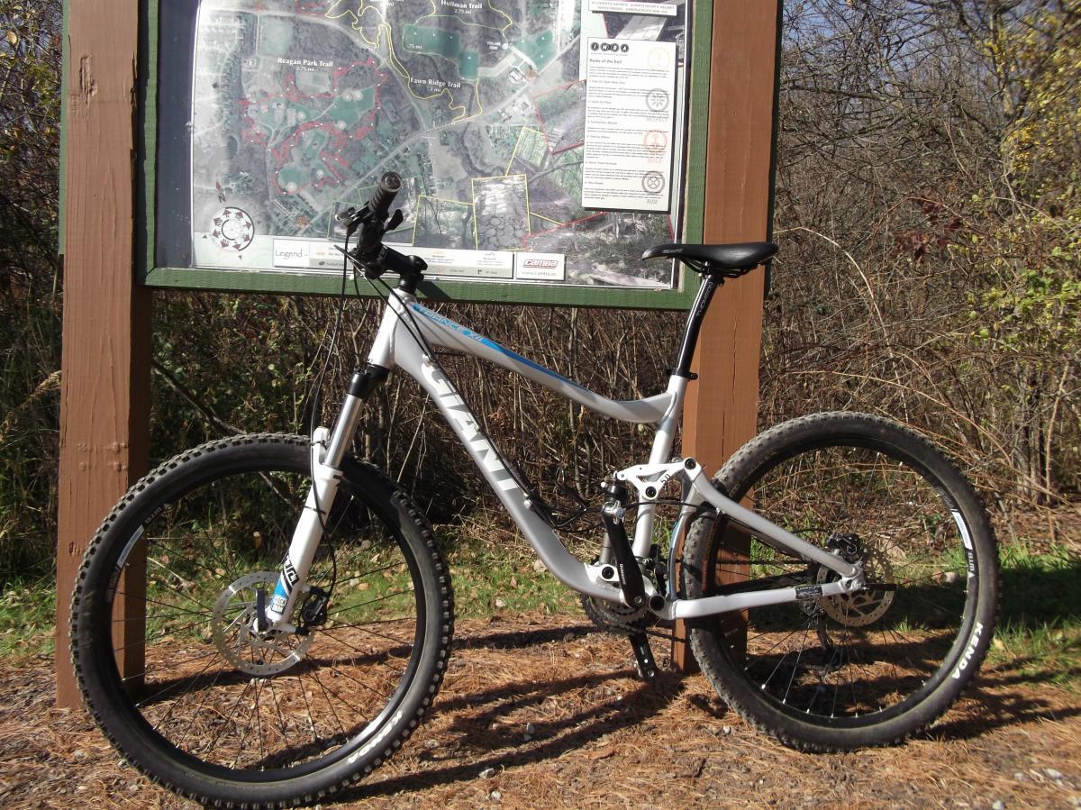 Giant Trance X4: A mountain bike parked beside an informational trail map displayed on a wooden sign. The bike has a light frame, thick tires, and a visible brand name. Surrounding the bike is a natural landscape with trees and underbrush. The map outlines various trails and features in the area.