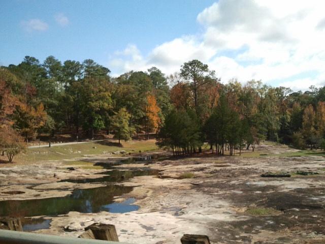 A scenic landscape featuring a calm, shallow stream surrounded by rocky terrain and a variety of trees. In the foreground, there are patches of exposed rock and water, while the background displays trees with autumn foliage, including shades of orange and green, under a partly cloudy blue sky. Flat Rock Park mountain bike trail.