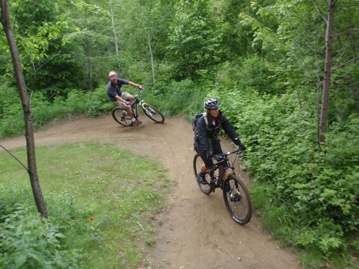 Two mountain bikers maneuvering a winding dirt trail through a lush green forest. One rider is leaning into a turn while wearing a red helmet and casual attire, and the other is approaching the turn wearing a black helmet and jacket. The surrounding area is filled with trees and vegetation, creating a natural biking environment. Kingdom Trails mountain bike trail.