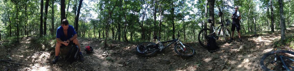 A panoramic view of a wooded area with two mountain bikers taking a break on a trail. One cyclist is seated on a rock, looking at a device, while the other stands nearby, holding a water bottle. Their mountain bikes are positioned on the ground next to them, surrounded by greenery and dirt paths. Coldwater Mountain mountain bike trail.