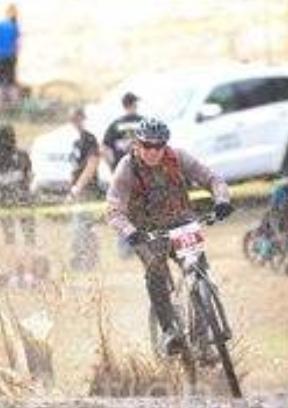 Trek Fuel EX 7: A mountain biker wearing a helmet and protective gear races on a dirt trail, splashing through a muddy section. The background features a crowd watching the event and a parked vehicle, indicating a competitive outdoor setting.
