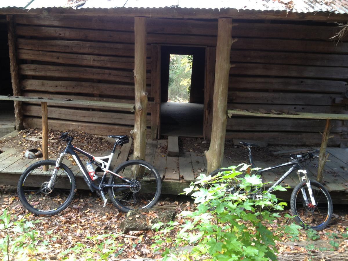 Two mountain bikes are parked next to a rustic wooden cabin in a forested area. The cabin has a weathered facade and an open doorway leading inside. The ground is covered with fallen leaves, and there are some green plants nearby. Clinton Nature Preserve mountain bike trail.