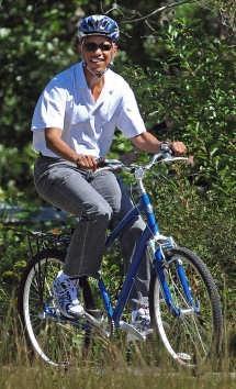 A person riding a blue bicycle on a sunny day, wearing a white shirt, gray pants, and a blue helmet, smiling while surrounded by greenery. Coldwater Mountain mountain bike trail.