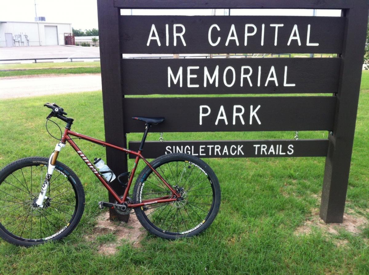 A mountain bike is positioned next to a sign for Air Capital Memorial Park, indicating the presence of singletrack trails. The park sign is painted in white lettering against a dark brown background and is surrounded by green grass. In the background, there is a glimpse of a building and road. Air Capital Memorial Park mountain bike trail.