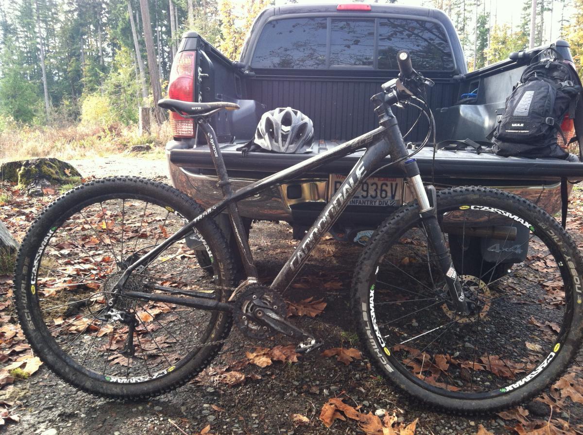 Cannondale Trail SL 29: A mountain bike is parked next to the back of a pickup truck in a wooded area, surrounded by fallen leaves. A gray helmet rests on the truck's tailgate, while a black backpack is visible on the truck bed. The scene is set in a natural environment with trees in the background, suggesting an outdoor adventure or biking trip.