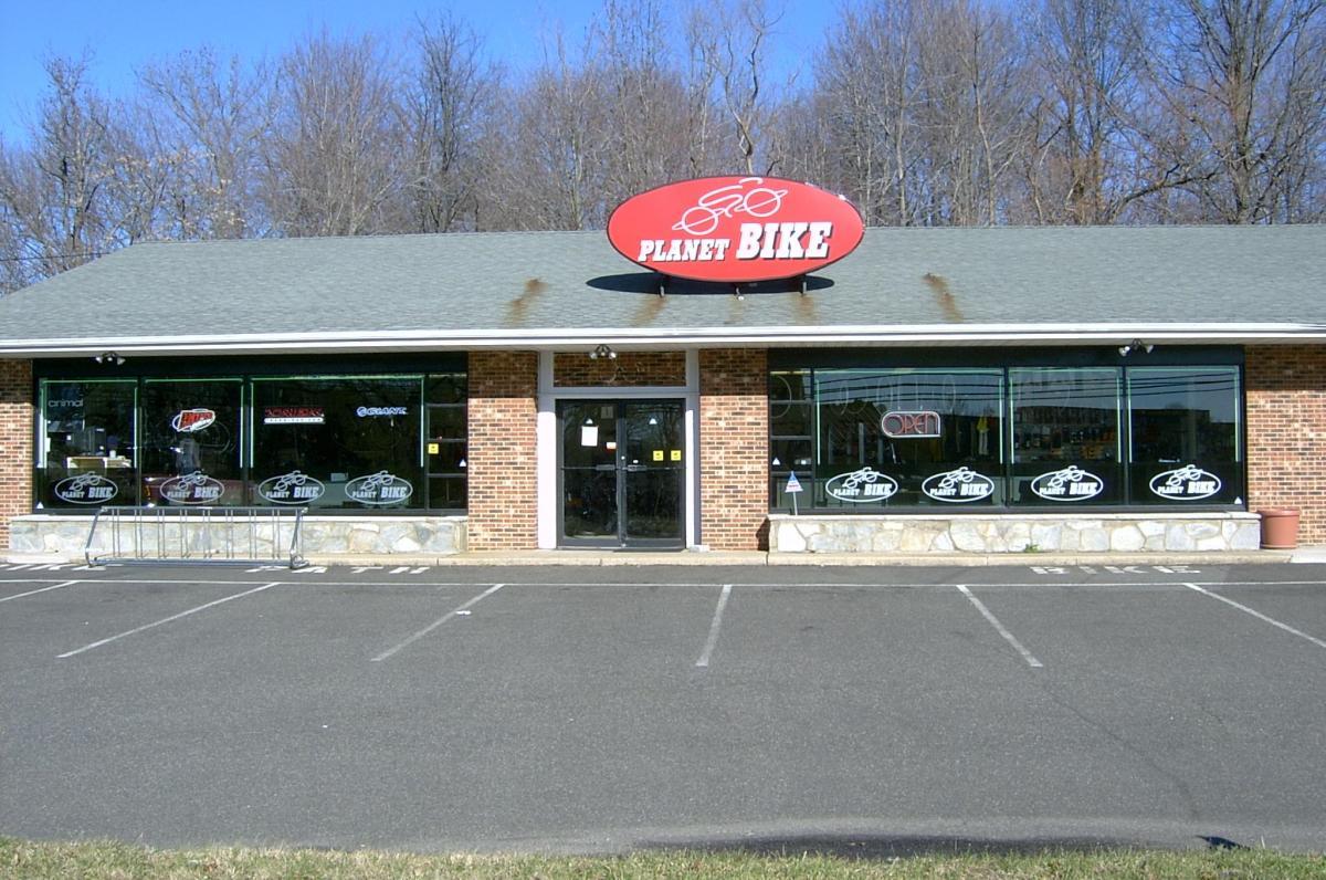 A brick building with a green roof and large windows showcasing bike-related merchandise, featuring a prominent red sign that reads "PLANET BIKE." The store has an "Open" sign visible and a bike rack in front, with several parking spaces in view. The background includes bare trees under a clear blue sky.