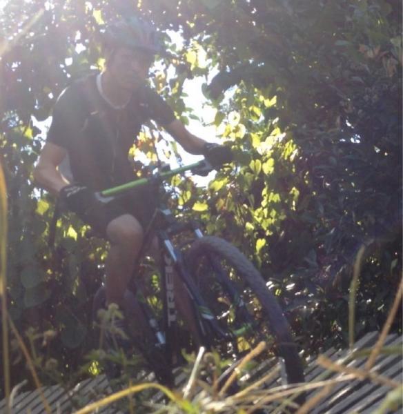 A mountain biker navigating a narrow, wooden trail through dense greenery, illuminated by sunlight filtering through the leaves. The biker is wearing a helmet and cycling gear, showing focus and control as they ride over the trail. Quiet Waters Park mountain bike trail.