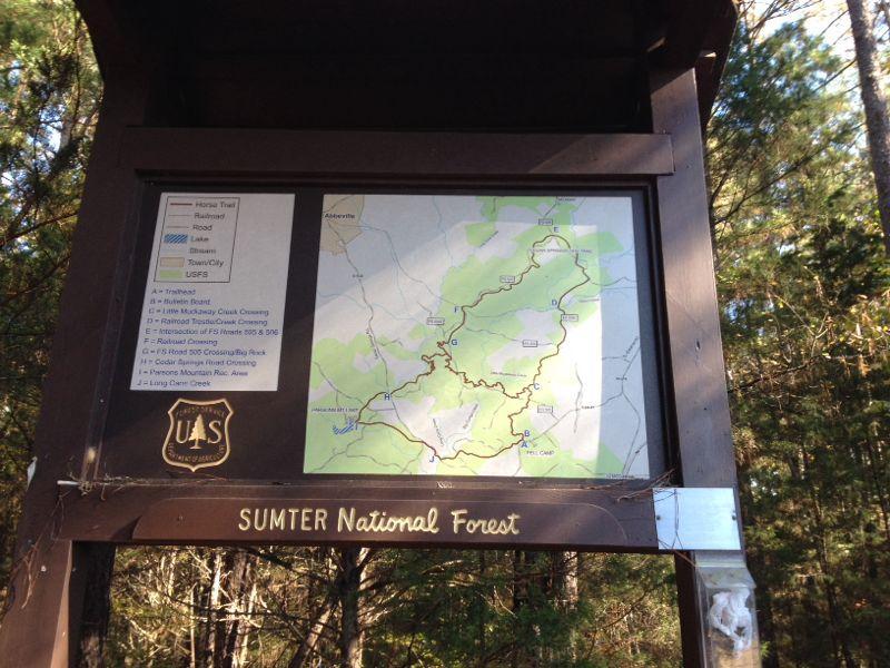 A sign displaying a detailed map of Sumter National Forest, with labeled trails, roads, and points of interest. The map includes a legend indicating different types of paths, such as horse trails and roads. Surrounding trees and greenery are visible in the background. Long Cane Horse Trail mountain bike trail.