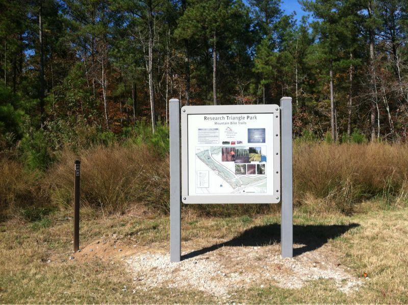 Signage for Research Triangle Park, detailing mountain bike trails, set against a backdrop of trees and tall grass. The sign includes images and maps of the trails. Research triangle park mountain bike trail.