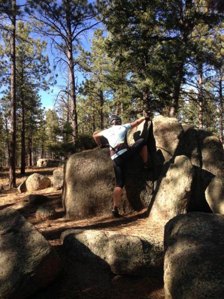 A climber in athletic attire is scaling a large rock in a forested area, surrounded by tall pine trees and boulders. The scene is set in natural light, showcasing a clear blue sky and a rugged outdoor environment. Buffalo Creek mountain bike trail.