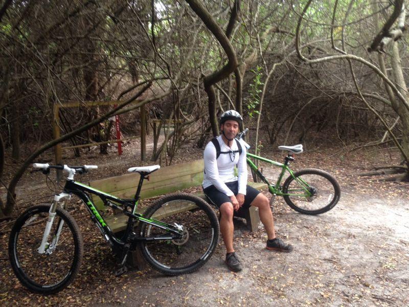 A person sitting on a wooden bench in a wooded area, wearing a helmet and athletic clothing. Two mountain bikes with green accents are placed beside the bench. The surroundings are filled with dense vegetation and trees. Caloosahatchee Regional Park mountain bike trail.