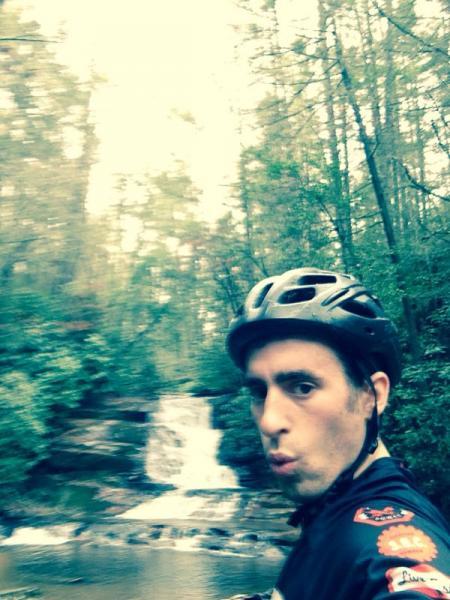 A cyclist wearing a helmet poses for a selfie in a wooded area, with a waterfall visible in the background. The scene is lush and green, capturing a moment of outdoor adventure. Stonewall Falls Loop mountain bike trail.