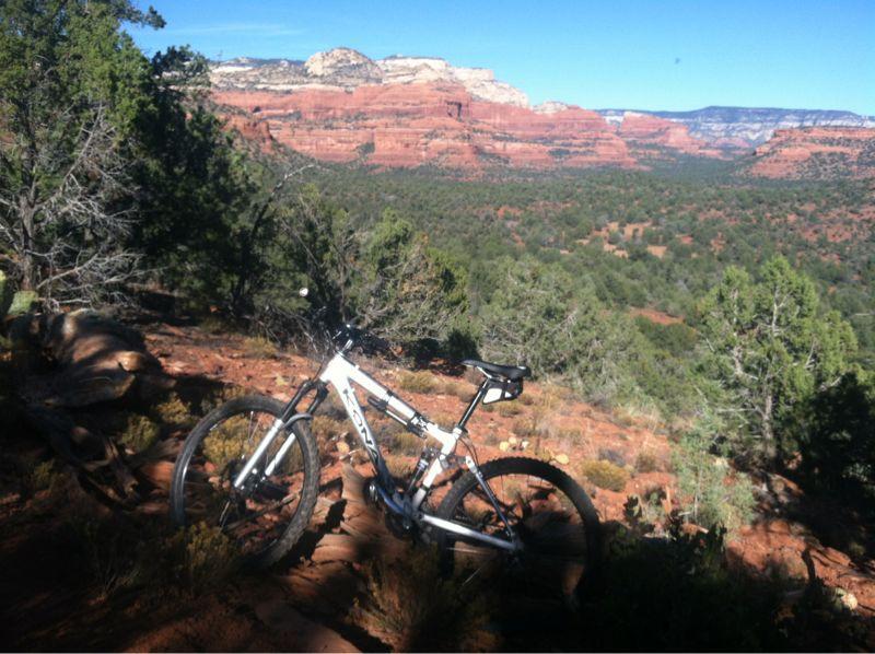 Alt tag: "A mountain bike parked on rocky terrain with a scenic view of red rock formations and lush green trees in the background." Long Canyon mountain bike trail.