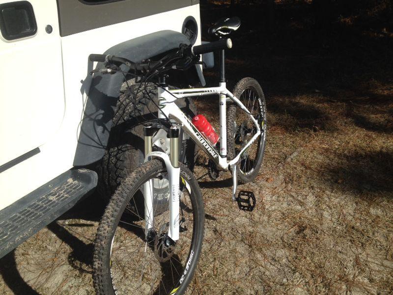 A white mountain bike leaning against a white vehicle, with a red water bottle attached to the frame. The scene is set in a natural environment, surrounded by pine needles and sparse vegetation. Bright sunlight is visible, casting shadows on the ground. Governor's Creek mountain bike trail.