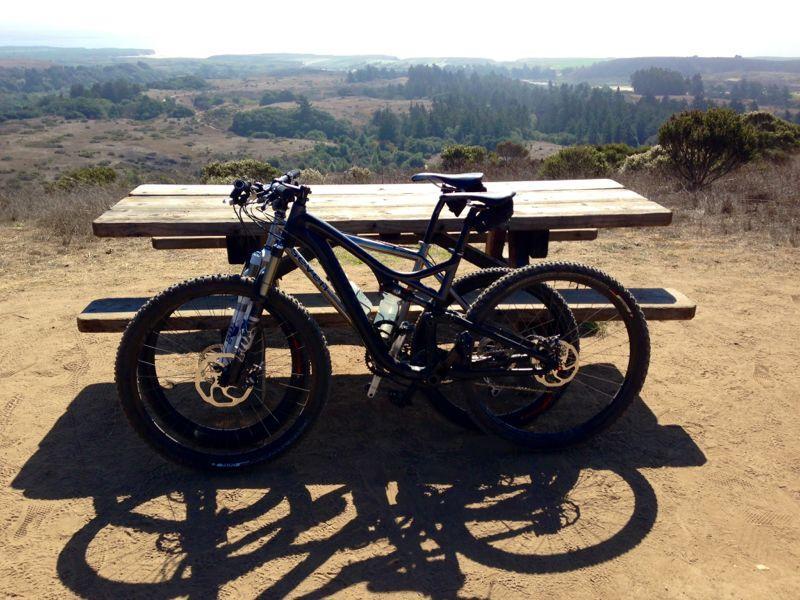 A mountain bike parked next to a wooden picnic table, with a scenic view of rolling hills and trees in the background under a clear sky. The bike's wheels cast shadows on the sandy ground. Wilder Ranch State Park mountain bike trail.