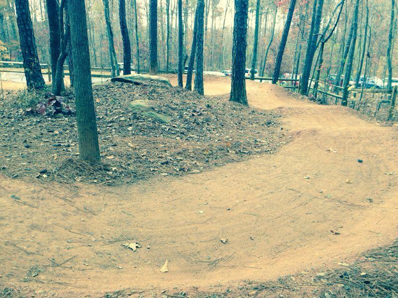 A dirt bike trail winding through a wooded area, with trees lining either side of the path and leaves scattered on the ground. In the background, parked cars and a wooden fence are visible. Big Creek mountain bike trail.