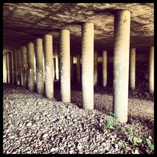 A view of a dimly lit underground space featuring several concrete columns supporting the ceiling. The ground is covered with loose gravel and stones, and there are hints of greenery emerging among the stones. The atmosphere is industrial and somewhat abandoned. Marian R. Bear Memorial Park mountain bike trail.