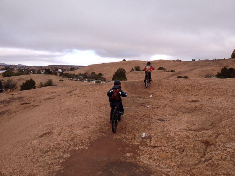 Two mountain bikers navigate a rocky trail on a cloudy day, with a landscape of hills and sparse vegetation in the background. Slickrock mountain bike trail.