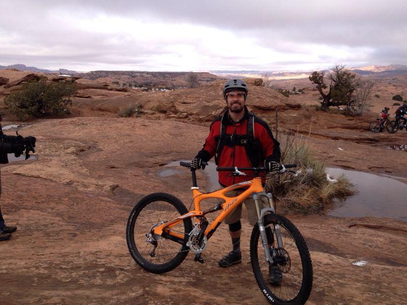 A man in a red jacket and helmet stands next to an orange mountain bike on rocky terrain. The landscape features a rugged background with shrubs and a cloudy sky, suggesting an outdoor cycling environment. Other cyclists are visible in the distance. Slickrock mountain bike trail.
