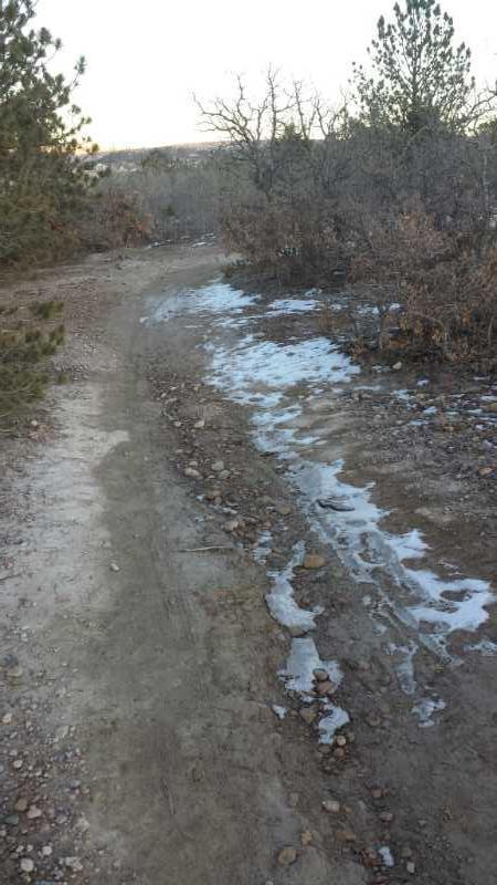 A winding dirt path through a forested area, bordered by sparse trees and brush. Sections of the path are covered with patches of snow and ice, indicating a cool climate. The landscape features rocky terrain and dry foliage, suggesting the changing seasons. Ute Valley Park mountain bike trail.