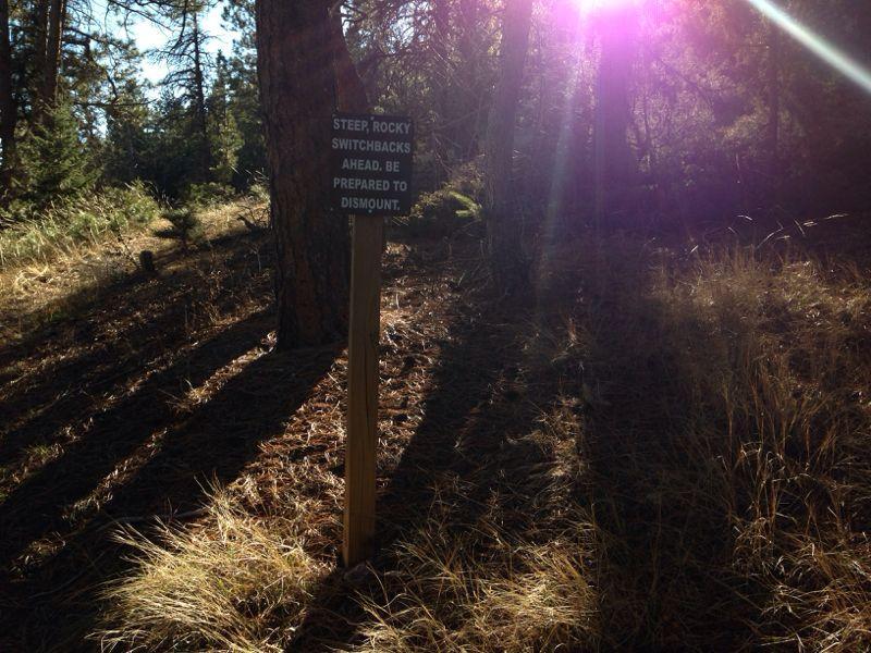 A sunlit forest area with tall trees and dry grass. A wooden sign is visible, reading "STEAP, ROCKY SWITCHBACKS AHEAD. BE PREPARED TO DISMOUNT," warning hikers of upcoming terrain difficulties. Lory State Park mountain bike trail.