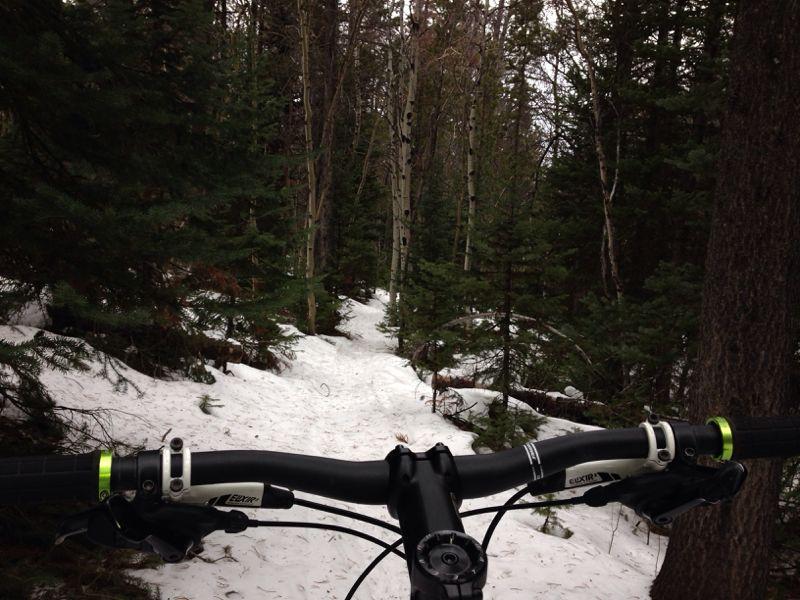 A view from the handlebars of a mountain bike, showcasing a snow-covered trail winding through a dense forest of tall trees and greenery. Happy Jack Recreation Area mountain bike trail.