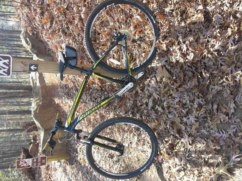 Mountain bike resting on a trail covered with autumn leaves, next to a wooden signpost indicating the direction for mountain biking. The bike features a black and yellow frame and is positioned in a wooded area, showcasing the natural surroundings. Fort Yargo State Park mountain bike trail.